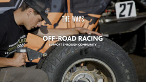 Person repairing large off-road tire with tool, text “THE WAYS OFF-ROAD RACING SUPPORT THROUGH COMMUNITY” and number 27 in background